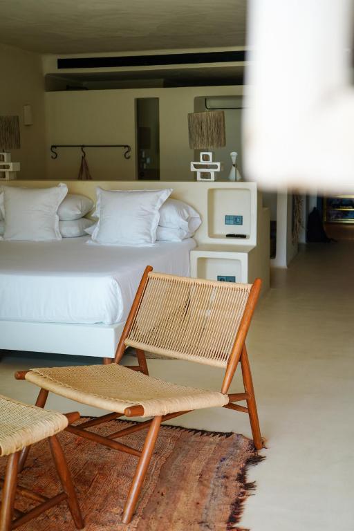 Modern minimalist hotel room with a neatly made bed, white linens, wooden furniture, a woven chair and footrest, and a rug on a light-colored floor—reflecting the simple, inviting style found in many boutique hotels in Formentera.