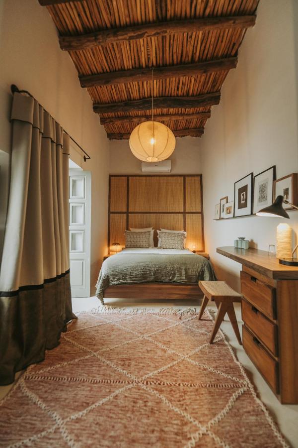 12 Charming Budget Riads in Marrakech 6 A cozy bedroom with a wooden beamed ceiling, a bed with green linens, and soft lighting—much like the inviting rooms found in budget riads in Marrakech—features a large woven rug, desk with lamp, and framed art for a warm atmosphere.