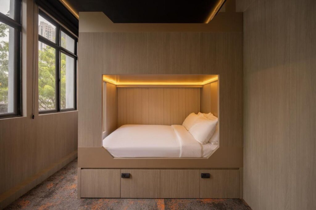13 Most Unique Hotels in Singapore for an Incredible Holiday 21 A modern, minimalistic bedroom inspired by unique hotels in Singapore, with a built-in wooden bed frame featuring soft lighting, white bedding and pillows. Large windows bring in natural light, and drawers are conveniently placed under the bed.