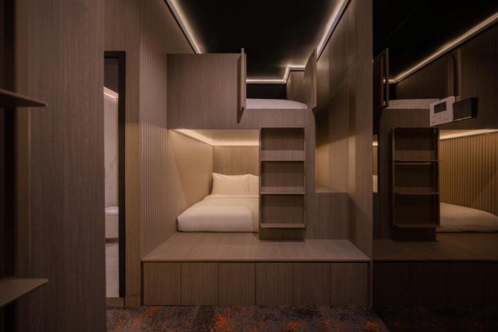 13 Most Unique Hotels in Singapore for an Incredible Holiday 22 A modern, minimalist room in one of the unique hotels in Singapore features two stacked wooden sleeping pods with cozy beds, soft lighting, a ladder, built-in shelves, neutral tones, and a bathroom visible through an open doorway.