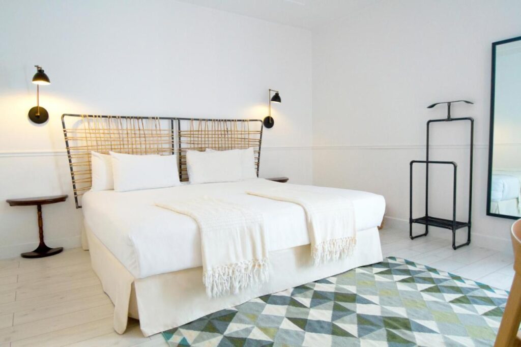 Where to Stay in Madrid: 14 Best Hotels 32 A bright, minimalist bedroom—perfect for deciding where to stay in Madrid first time—with a large bed, woven headboard, white bedding, wall-mounted lamps, small round side tables, a green geometric rug, tall mirror, and black valet stand.