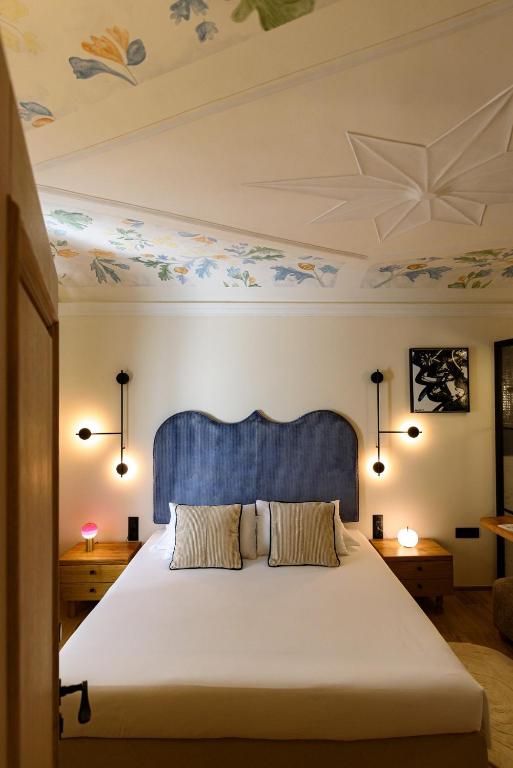 Most Unique Boutique Hotels in Dubrovnik 14 A cozy bedroom inspired by boutique hotels in Dubrovnik, with a bed featuring a blue upholstered headboard, striped pillows, wooden nightstands with lamps, modern wall lights, and a ceiling adorned with floral patterns and a star motif.