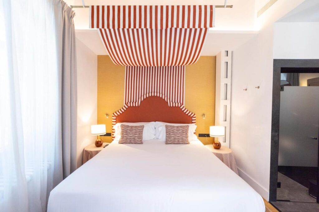 10 UNIQUE Hotels with Pool in Seville 11 A neatly made bed with a white duvet and two striped pillows stands beneath a red and white striped canopy—evoking the charming style found in many hotels with pool in Seville. Two bedside tables with lamps flank the bed, beside a large window with sheer curtains.