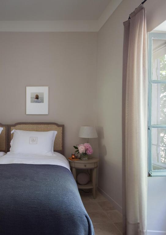 11 Unique Places to Stay in Provence 42 A neatly made bed with a blue blanket and white pillows sits beside a small round nightstand holding flowers, a book, and fruit. Light filters in through an open window—perfect for those seeking unique places to stay in Provence.