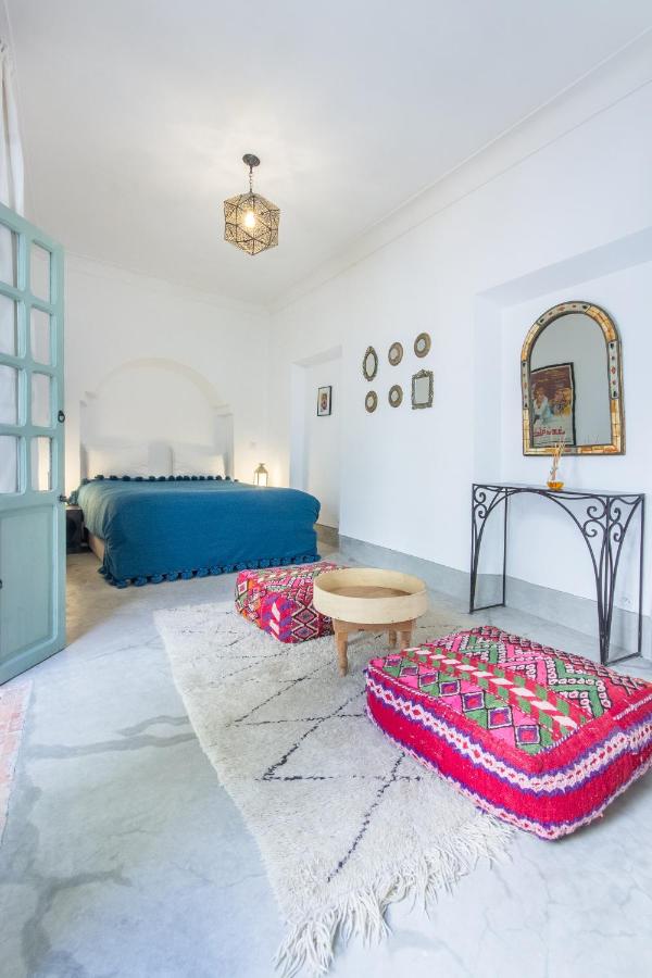 12 Charming Budget Riads in Marrakech 18 A bright, airy bedroom inspired by budget riads in Marrakech, with white walls, a blue bedspread, patterned floor cushions, a round wooden table, a rug, wall decor, a mirror on a metal console, and a geometric light fixture.