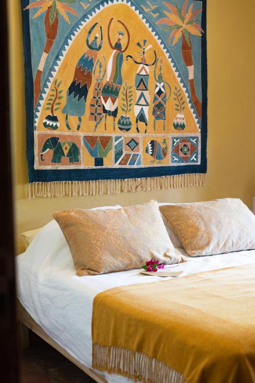 Adults-Only Hotels in Costa del Sol 29 A cozy bed with two tan pillows and a yellow blanket, topped with a small plate of flowers, sits beneath a vibrant tapestry of figures and palm trees—perfect inspiration for adult only hotels in Costa del Sol.