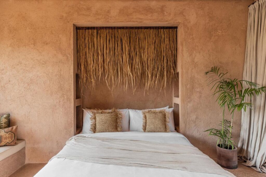 A cozy bedroom in one of the best family villas in Bali, featuring a bed against a textured beige wall, straw decor above the headboard, woven pillows, neutral bedding, a potted plant, and light curtains for an earthy atmosphere.