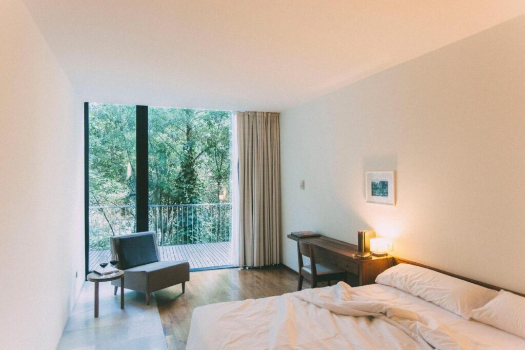 4 Best Hotels in Ribera del Duero, Spain 15 Minimalist bedroom with a large window overlooking trees, perfect for planning weekend trips from Madrid. Features include a bed with white bedding, a wooden desk and lamp, a chair by the window, and light curtains. Natural light fills the space.