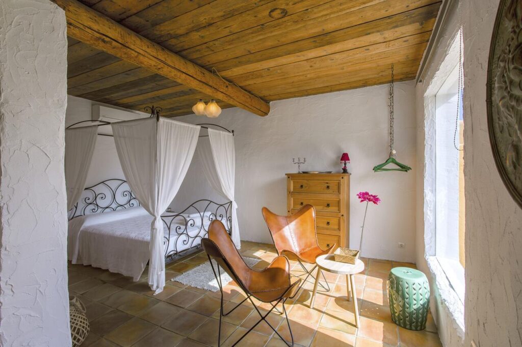 10 AUTHENTIC Finca Hotels in Ibiza 16 Sunny bedroom inspired by finca hotels in Ibiza, featuring a wooden ceiling, canopy bed with white curtains, wooden dresser, two leather chairs, small table, green stool, and a large window letting in natural light.