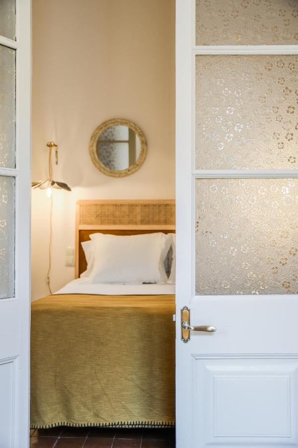 A cozy bedroom, reminiscent of boutique hotels in Costa Brava, is seen through a partially open white door, featuring a neatly made bed with white pillows and a mustard yellow blanket, a round mirror above the headboard, and a wall-mounted lamp.