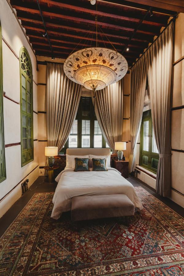 9 Best Hotels in Saudi Arabia: Unique Stays Across the Country 2 A cozy bedroom like one in the Best Hotels in Saudi Arabia, with a large patterned rug, a bed with white linens and decorative pillows, two bedside tables with lamps, tall draped curtains, green-framed windows, and an ornate ceiling light.