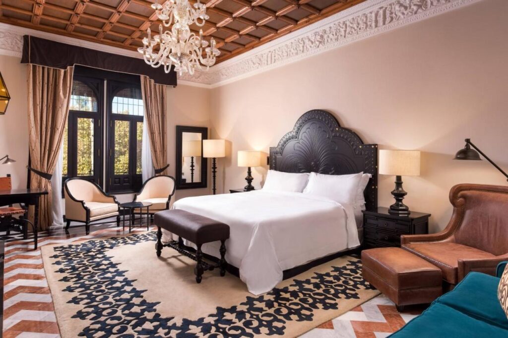 10 UNIQUE Hotels with Pool in Seville 23 Elegant hotel room with a large bed, ornate black headboard, chandelier, patterned rug, and armchairs near tall windows. Enjoy classic and modern furnishings in warm tones at one of the top hotels with pool in Seville.