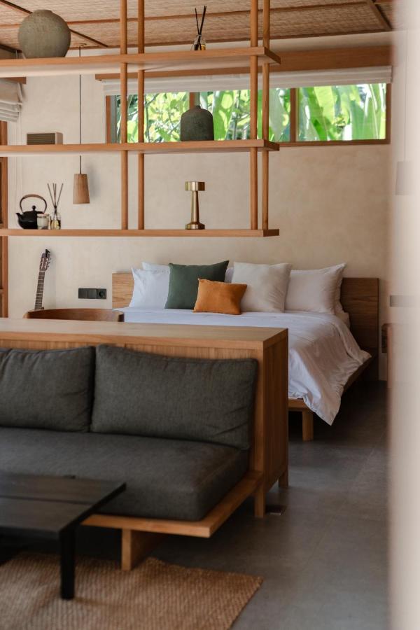 Modern, minimalist bedroom in one of the best family villas in Bali, featuring a wooden bed, white and green bedding, small sofa, black coffee table, open shelving, and large windows with views of lush greenery.