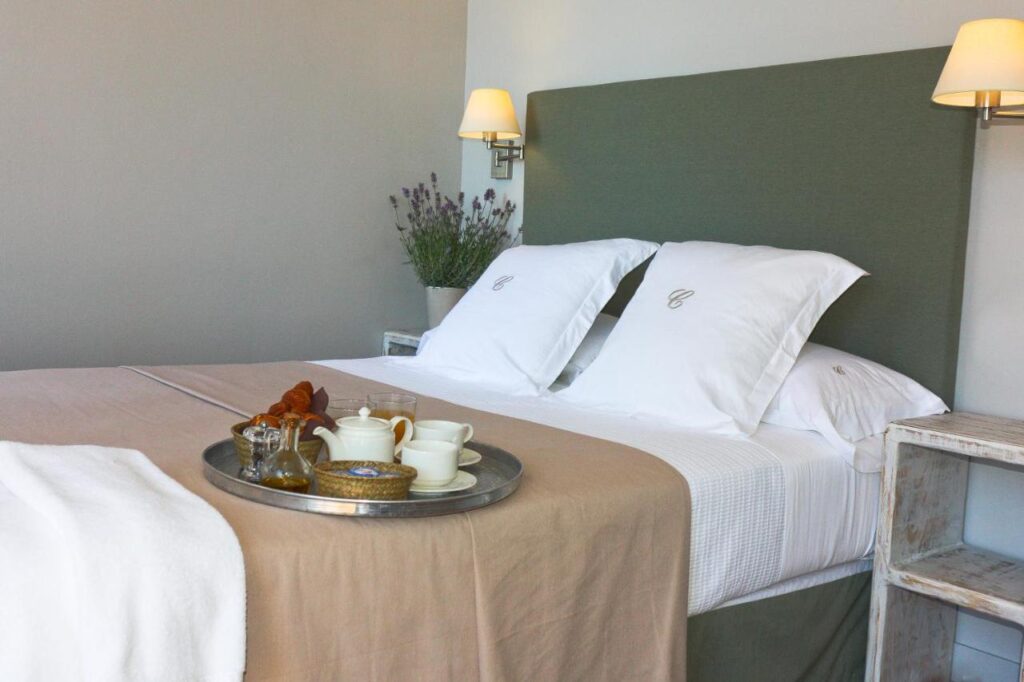 A neatly made bed with white and beige linens, monogrammed pillows, and a green headboard evokes the charm of boutique hotels in Costa Brava. A tray with tea sits atop the bed, flanked by bedside tables, wall lamps, and a vase of lavender.