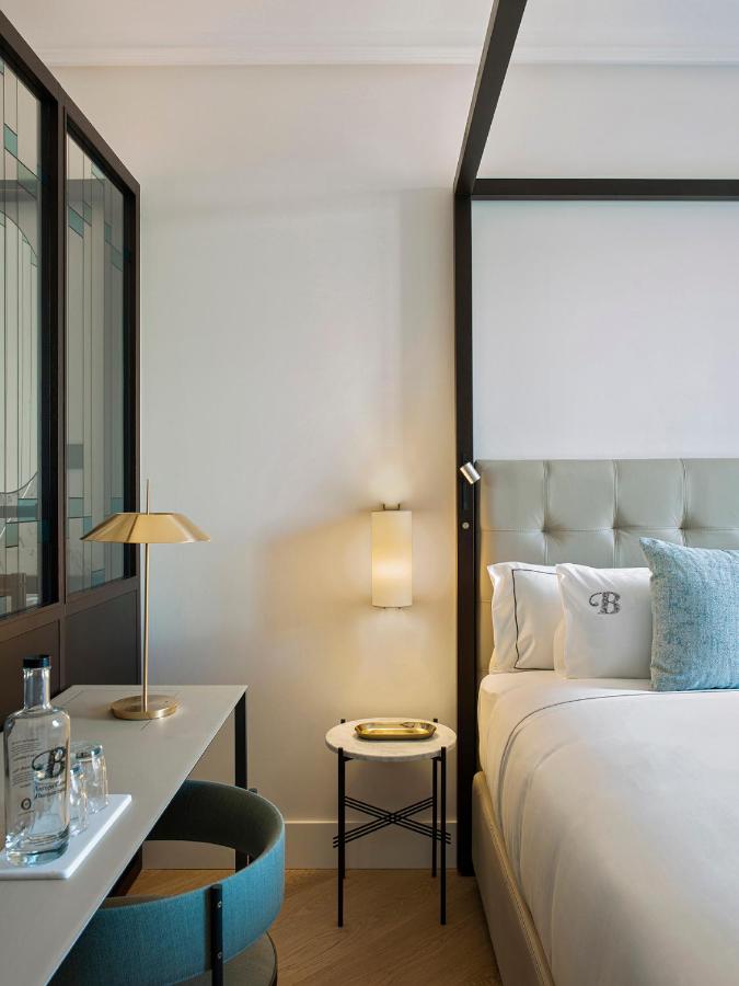 Top Barcelona Hotels with Rooftop Pool 7 A modern, minimalistic hotel room featuring a tufted headboard, decorative pillows, a side table, desk with lamp, chair, and a bottle of water. Neutral and light blue tones echo the style of top Barcelona hotels with rooftop pools.