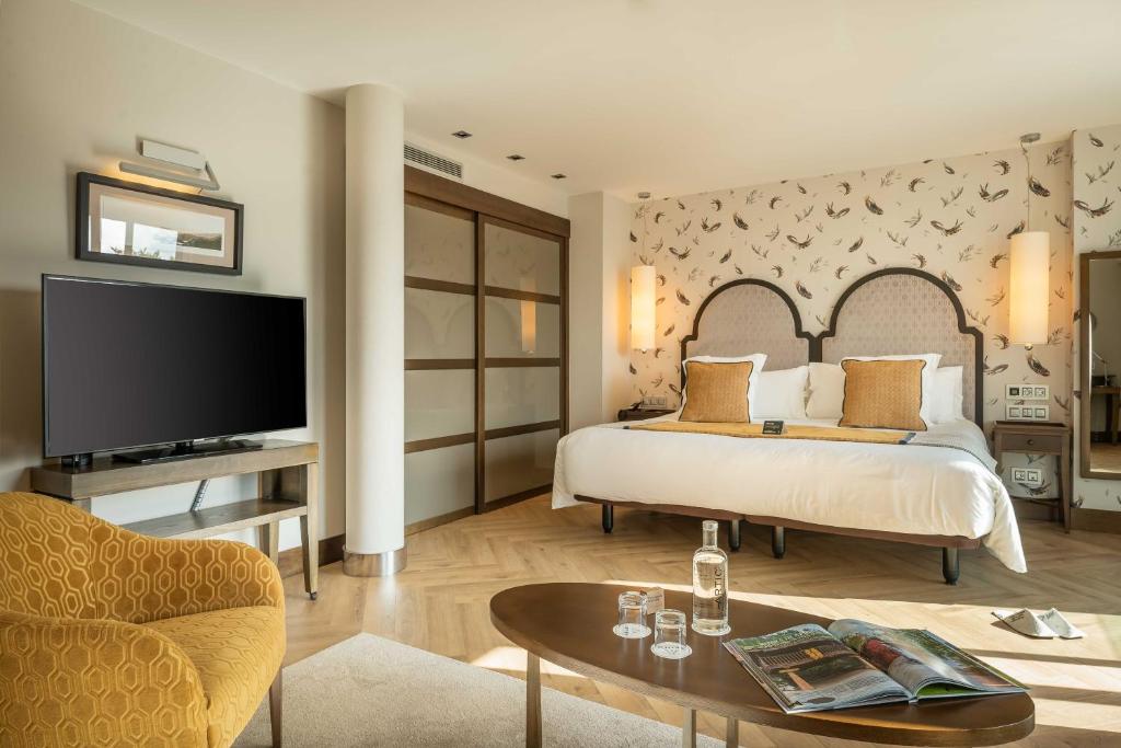 A stylish boutique hotel room in Asturias with a large bed, two yellow cushions, patterned wallpaper, a flat-screen TV, a yellow chair, a modern coffee table with glasses and a water bottle, and warm ambient lighting.