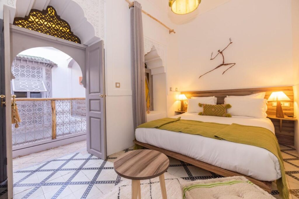 12 Charming Budget Riads in Marrakech 26 A bright, cozy bedroom in one of the charming budget riads in Marrakech, with a large bed covered in white linens and a green throw. Warm lamps light the room, which features arched doorways, tile floors, and an open door leading to a balcony.