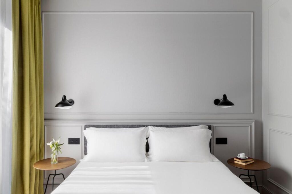 Where to Stay in Madrid: 14 Best Hotels 52 A modern, minimalistic bedroom ideal for those seeking where to stay in Madrid first time, featuring a neatly made bed with white linens, black reading lamps, small wooden side tables, fresh flowers, books, and green curtains by the window.