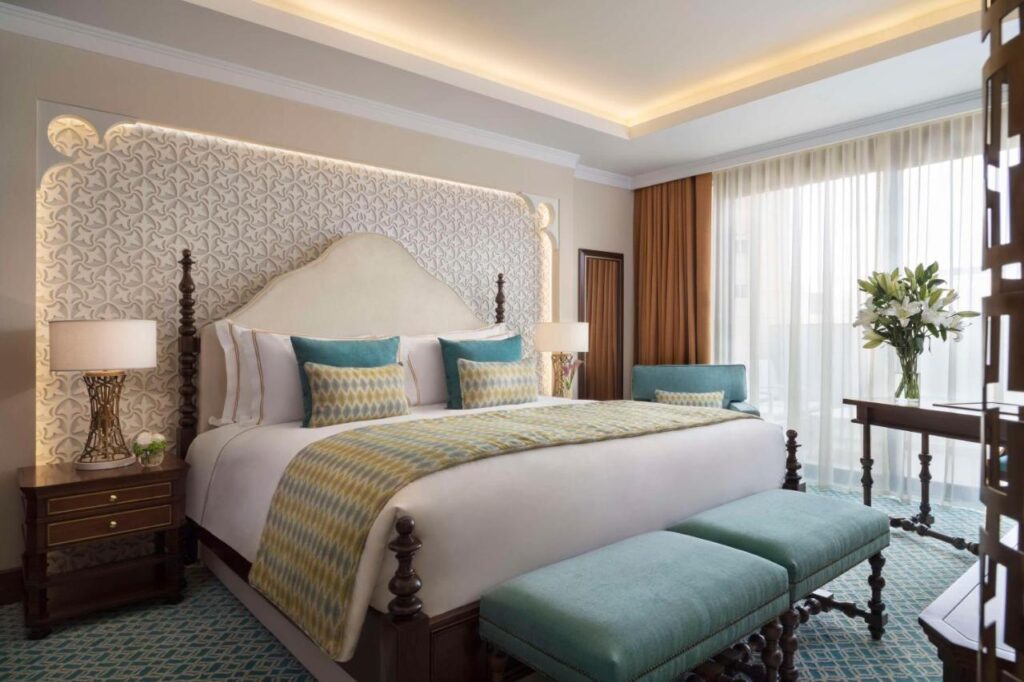 Where to Stay in Doha, Qatar: Transit, City & Beach Hotels 43 A luxurious hotel room—ideal for those seeking where to stay in Doha—features a large bed with an elegant headboard, plush pillows, patterned bedding, two bedside tables with lamps, a blue bench, fresh flowers, and floor-to-ceiling windows with sheer curtains.