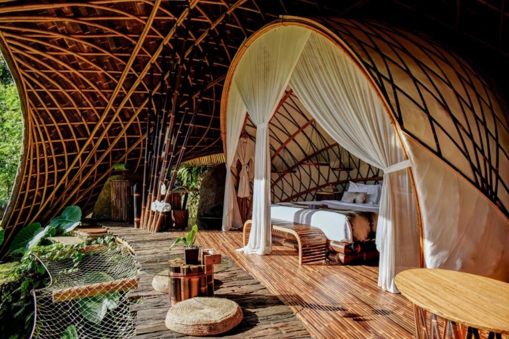 13 Most UNIQUE Places to Stay in Ubud 13 A luxurious bamboo and canvas eco-lodge, one of the unique places to stay in Ubud, features a cozy bed with white curtains, wooden furniture, and large windows opening to lush greenery, blending modern comfort with nature.