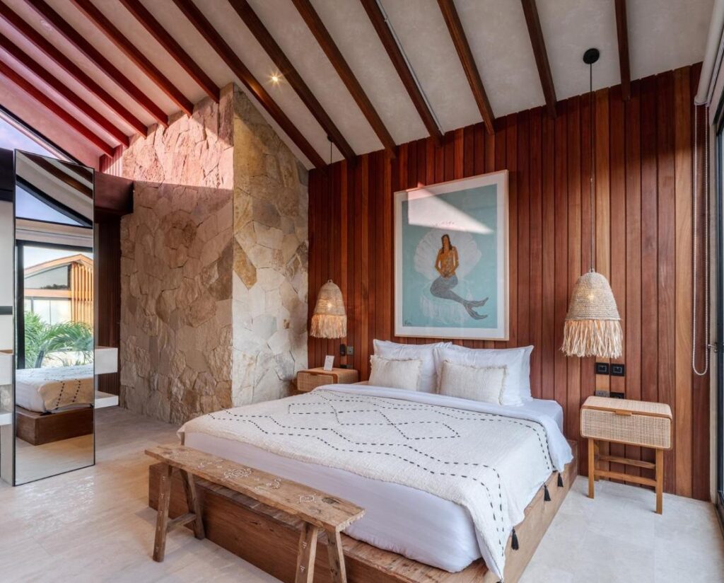 14 UNIQUE Boutique Hotels in Canggu, Bali 54 A modern bedroom inspired by boutique hotels in Canggu, with wood-paneled walls, a large bed with white bedding, a wooden bench at the foot, woven lamps, and wall art. Natural light pours through large windows and highlights the stone accent wall.
