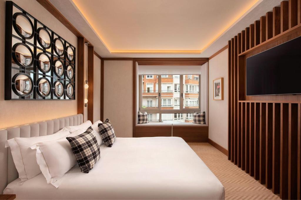 8 Unique Boutique Hotels in Bilbao Spain 14 A modern hotel room at one of the boutique hotels in Bilbao features a large bed, plaid pillows, a wall-mounted TV, circular mirrors above the headboard, and a window seat with cushions overlooking city buildings. Warm lighting highlights wooden accents.