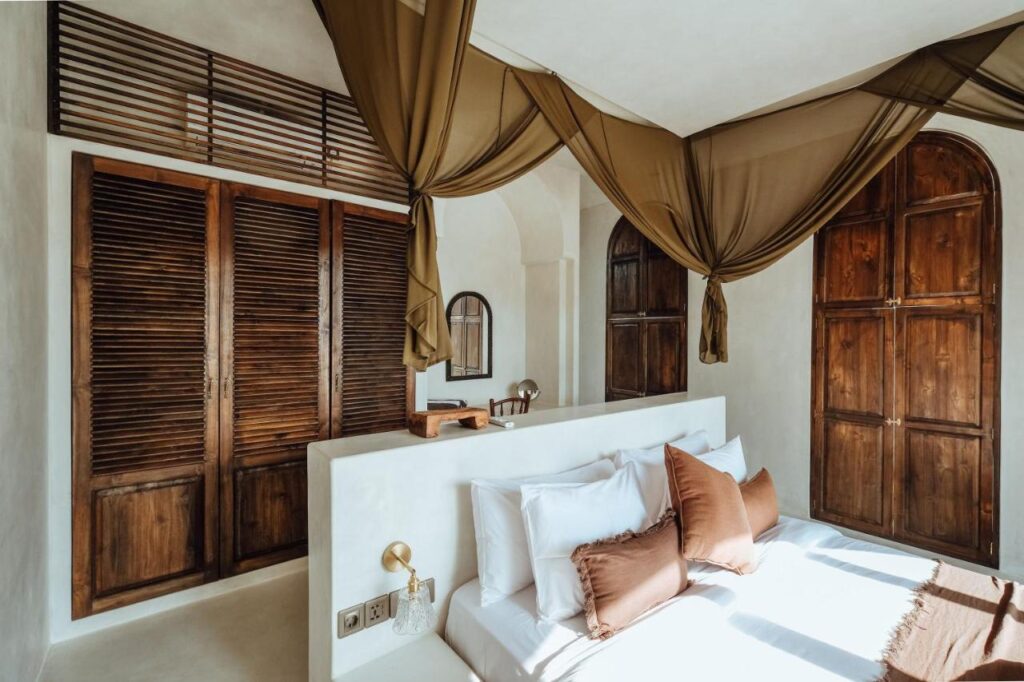 14 UNIQUE Boutique Hotels in Canggu, Bali 17 A sunlit bedroom with white walls, brown wooden shutters, sheer draped canopy, and a bed with white linens and brown pillows—perfectly echoing the serene style found in boutique hotels in Canggu. Gold wall lamps and a round mirror add elegant touches above the desk.