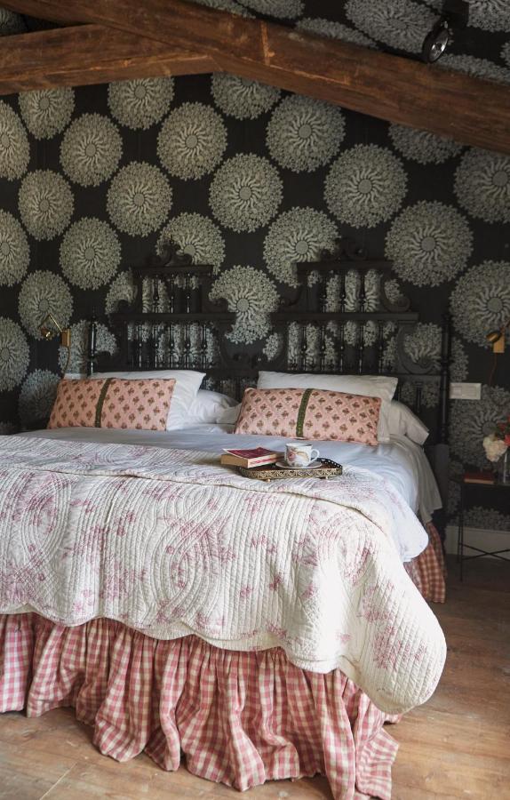 25 Weekend Trips from Madrid Max. 2h Away 34 A cozy bedroom with a large bed covered in a white and pink quilt and checkered bed skirt—perfect for relaxing after weekend trips from Madrid. Two pillows with pink covers, a cup, books on the bed, and floral wallpaper create a charming retreat.