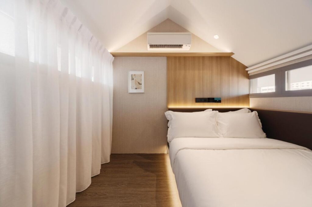 13 Most Unique Hotels in Singapore for an Incredible Holiday 27 A small, modern bedroom at one of the unique hotels in Singapore, featuring a double bed with white bedding, wooden floor, wood-paneled wall, air conditioner above the bed, framed picture, and sheer white curtains covering a window on the left.