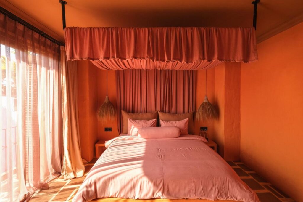 14 UNIQUE Boutique Hotels in Canggu, Bali 46 A cozy bedroom with pink bedding, a pink canopy, and stylish hanging lights—reminiscent of boutique hotels in Canggu. Warm sunlight streams through sheer curtains, illuminating the orange-toned walls and creating a soft, inviting atmosphere.