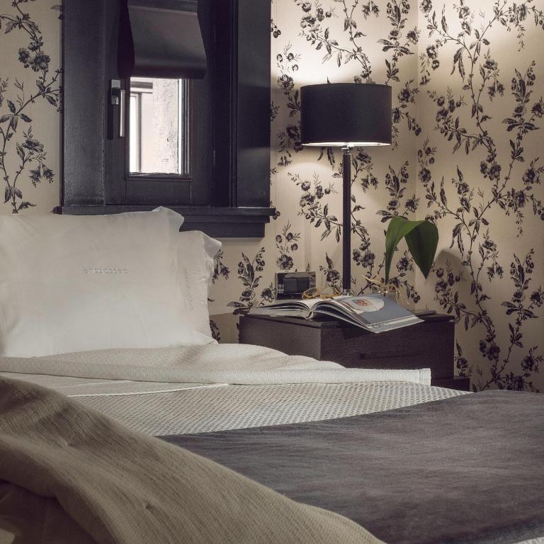 8 Unique Boutique Hotels in Bilbao Spain 32 A cozy bedroom reminiscent of Boutique Hotels in Bilbao, with a white pillow and blanket on the bed, a bedside table holding books, a lamp, a plant, and a clock, set against floral-patterned wallpaper and a window in the background.