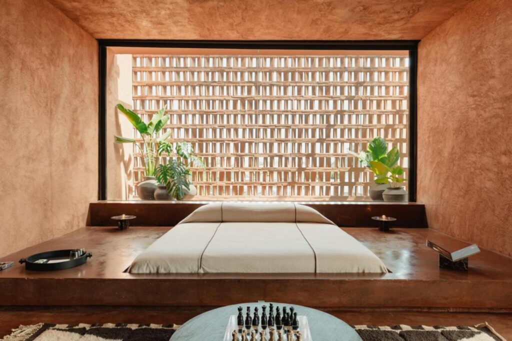 14 UNIQUE Boutique Hotels in Canggu, Bali 10 A minimalist bedroom inspired by boutique hotels in Canggu, with a low, built-in bed, earth-toned walls, potted plants, and a geometric brick screen window. A chess set and modern decor accent the serene, sunlit space.