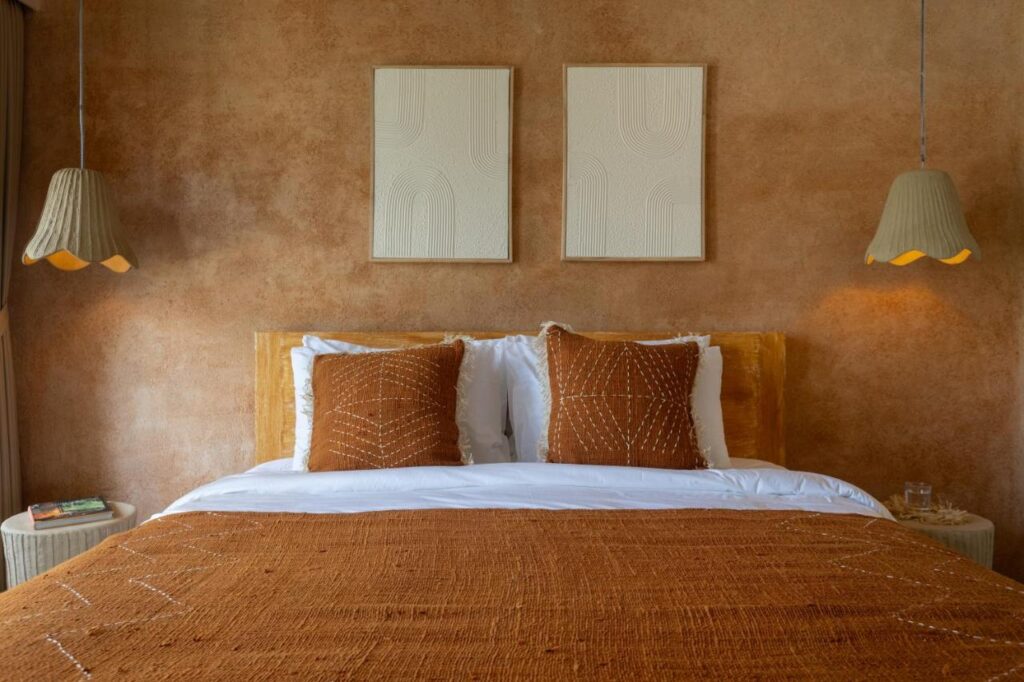 14 UNIQUE Boutique Hotels in Canggu, Bali 34 A cozy bedroom inspired by boutique hotels in Canggu, with a bed featuring white sheets, a rust-colored blanket, and matching pillows. Two hanging pendant lights and minimalist art decorate the textured brown wall above the bed.