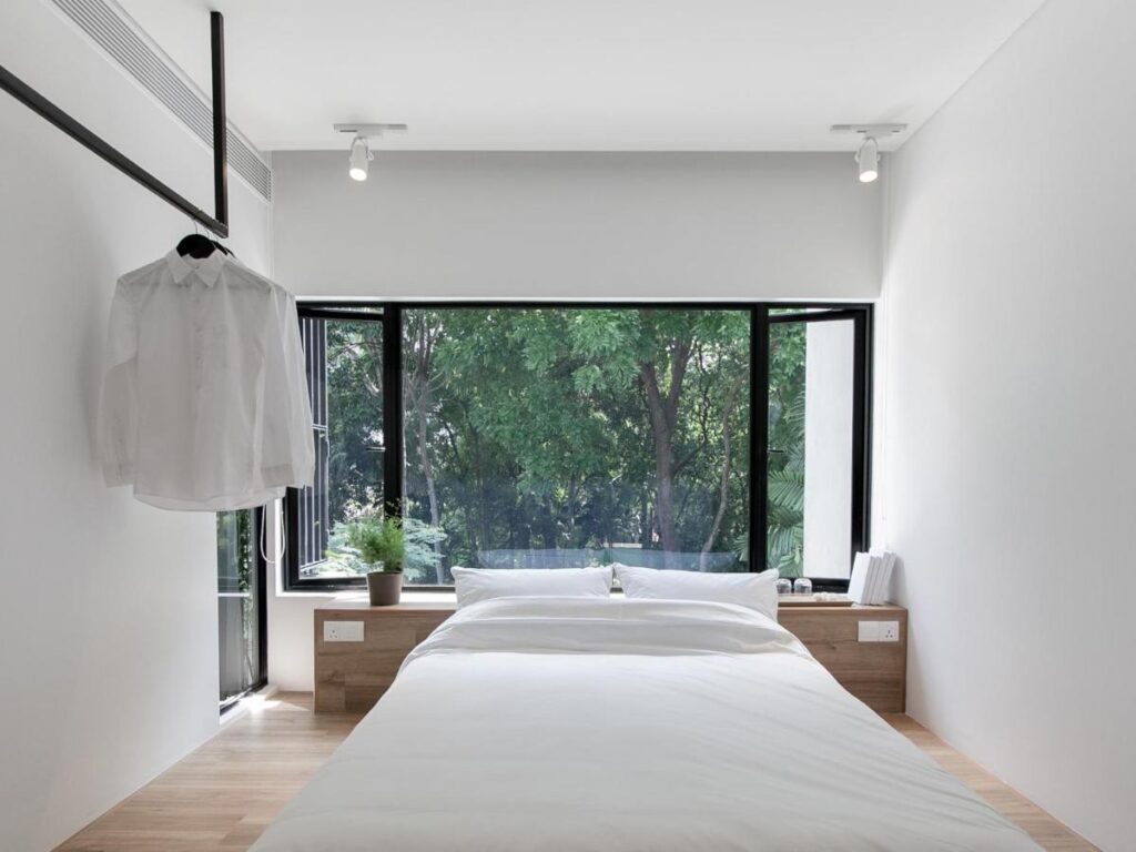 13 Most Unique Hotels in Singapore for an Incredible Holiday 12 A minimalist bedroom with a large window overlooking trees, a neatly made bed with white linens, wooden bedside tables, and a white shirt hanging on a rail by the window—natural light fills this space, reminiscent of unique hotels in Singapore.