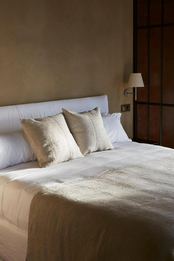Best Boutique Hotels in San Sebastian 20 A neatly made bed with light beige bedding and two matching pillows, reminiscent of boutique hotels in San Sebastian, sits against a neutral wall with a wall-mounted lamp and a window with black frames. Soft natural light fills the room.