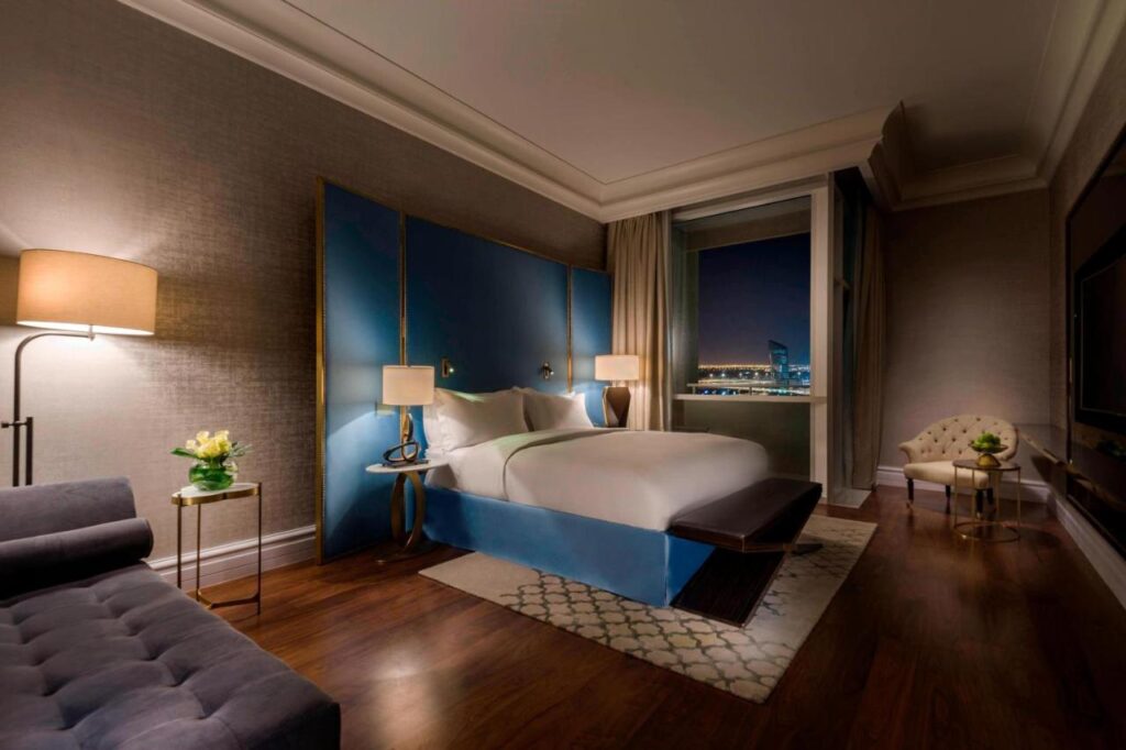 Where to Stay in Doha, Qatar: Transit, City & Beach Hotels 19 Modern hotel room with a large bed, blue headboard, two bedside lamps, sofa, armchair, and floor lamp. Large window offers a cityscape view—ideal for travelers searching where to stay in Doha with comfort and style.