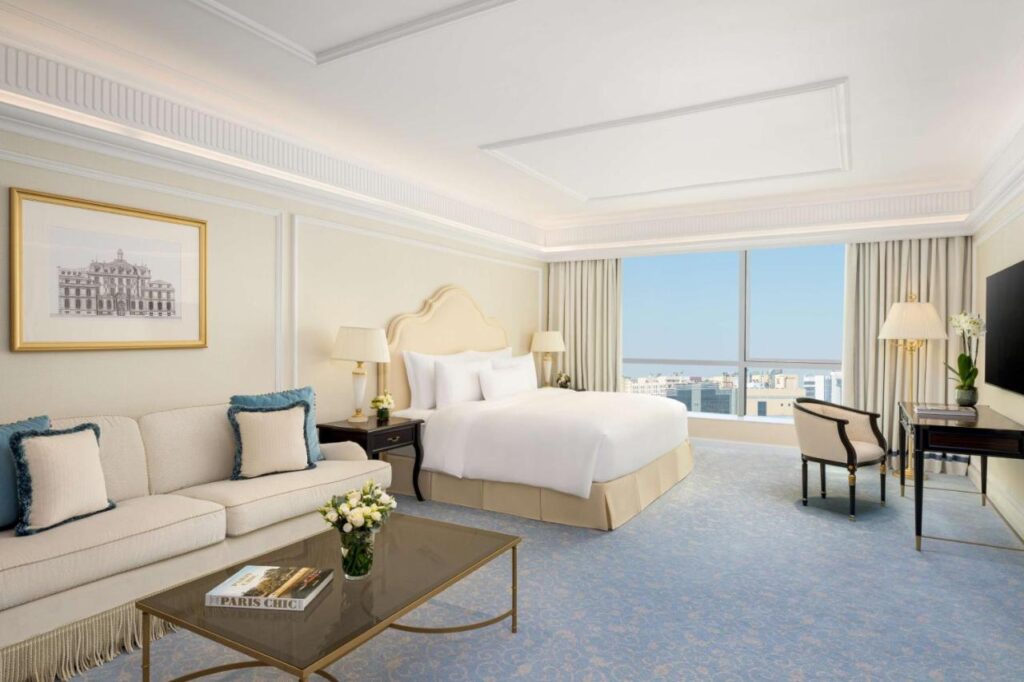 Where to Stay in Doha, Qatar: Transit, City & Beach Hotels 30 Elegant hotel room with a large bed, cream-colored walls, sofa and pillows, glass coffee table with flowers—an ideal choice for those seeking where to stay in Doha. Enjoy a desk, flat-screen TV, city views from a large window, and a soft blue carpet.