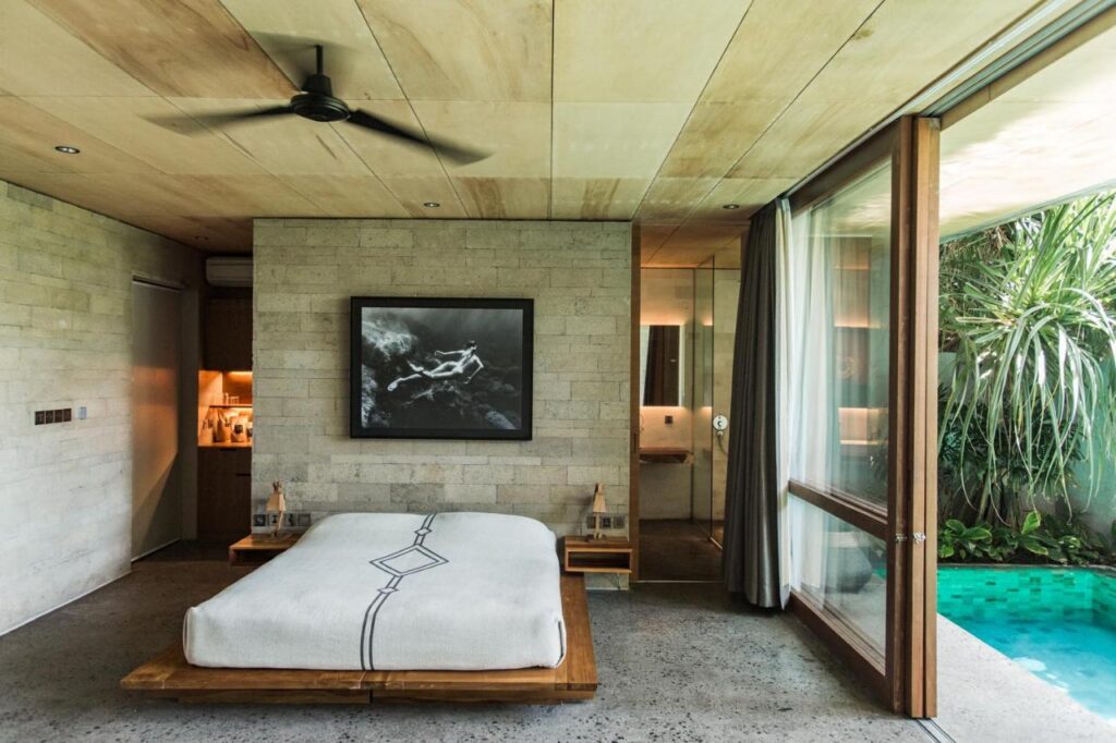 14 UNIQUE Boutique Hotels in Canggu, Bali 22 A modern bedroom inspired by boutique hotels in Canggu, with a minimalist bed, concrete walls, a ceiling fan, large glass doors overlooking a private pool and lush greenery. A black-and-white photo hangs above the bed.