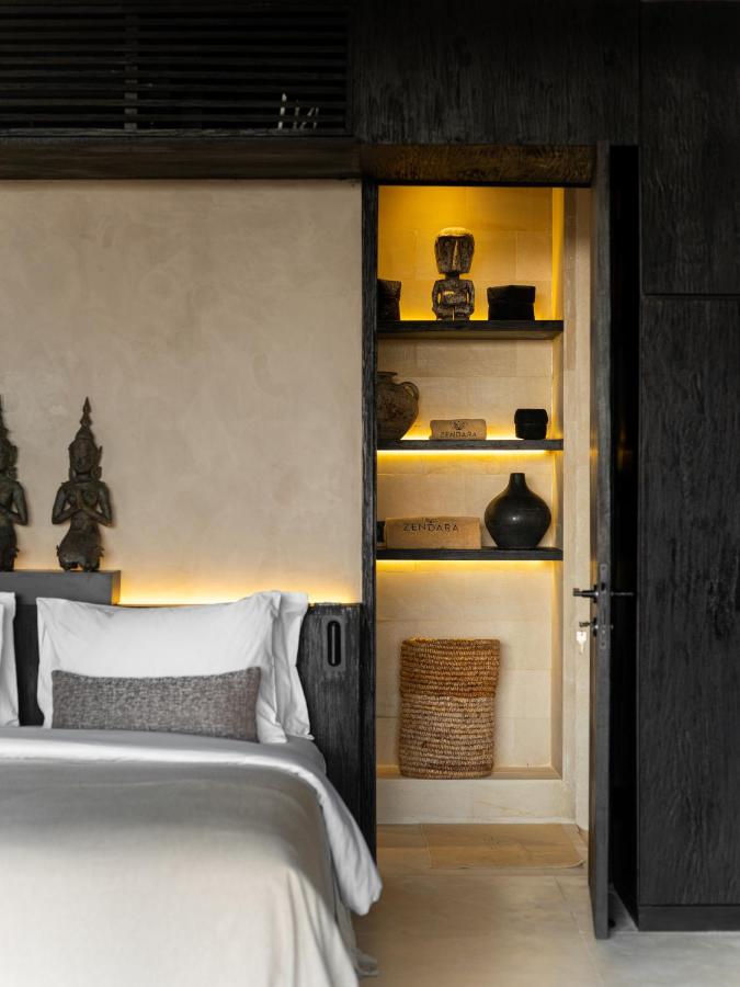14 UNIQUE Boutique Hotels in Canggu, Bali 51 A modern bedroom with a bed featuring white linens and gray accents, reminiscent of boutique hotels in Canggu, beside a built-in alcove with shelves displaying decorative vases, sculptures, a woven basket, and warm ambient lighting.