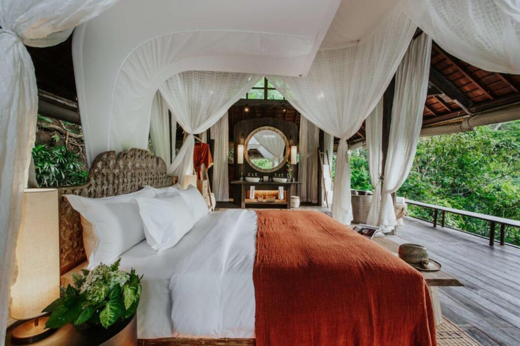 13 Most UNIQUE Places to Stay in Ubud 14 A luxurious canopy bed with white linens and a rust-colored blanket sits in an open-air wooden villa, surrounded by lush greenery—one of the most unique places to stay in Ubud, with elegant decor and an inviting outdoor deck.