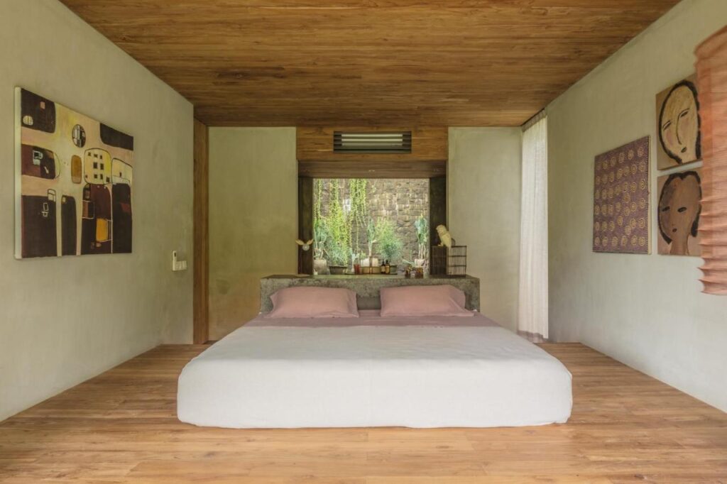 A minimalist bedroom with a low platform bed, light bedding, wooden floor and ceiling, abstract art on the walls, and a large window behind the bed overlooking greenery—evoking the serene style of the best family villas in Bali.