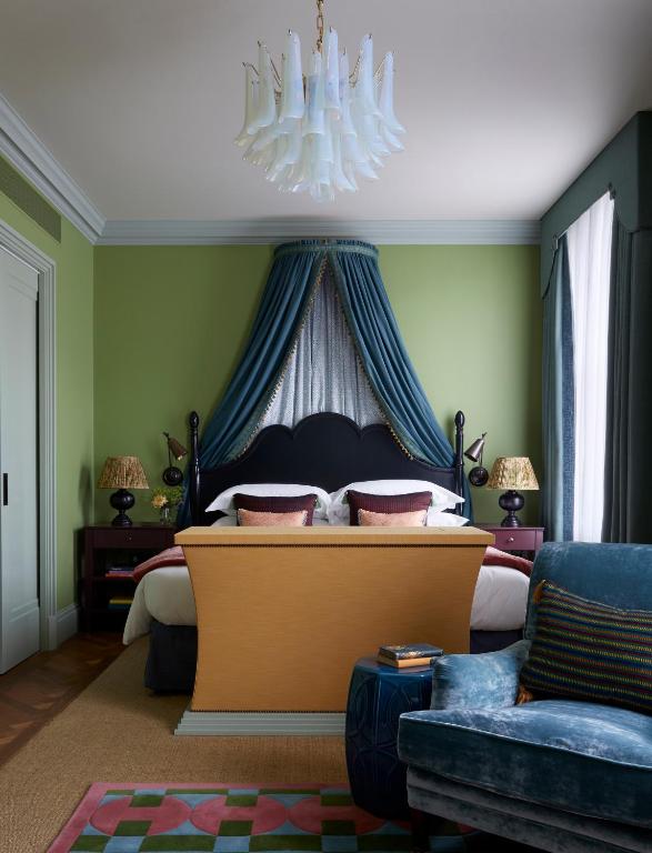 15 Best Boutique Hotels in London, UK 46 A stylish bedroom inspired by boutique hotels in London, with green walls, a black bed frame, blue drapes above the bed, patterned pillows, a blue velvet armchair, a modern chandelier, and natural light from tall windows with white curtains.