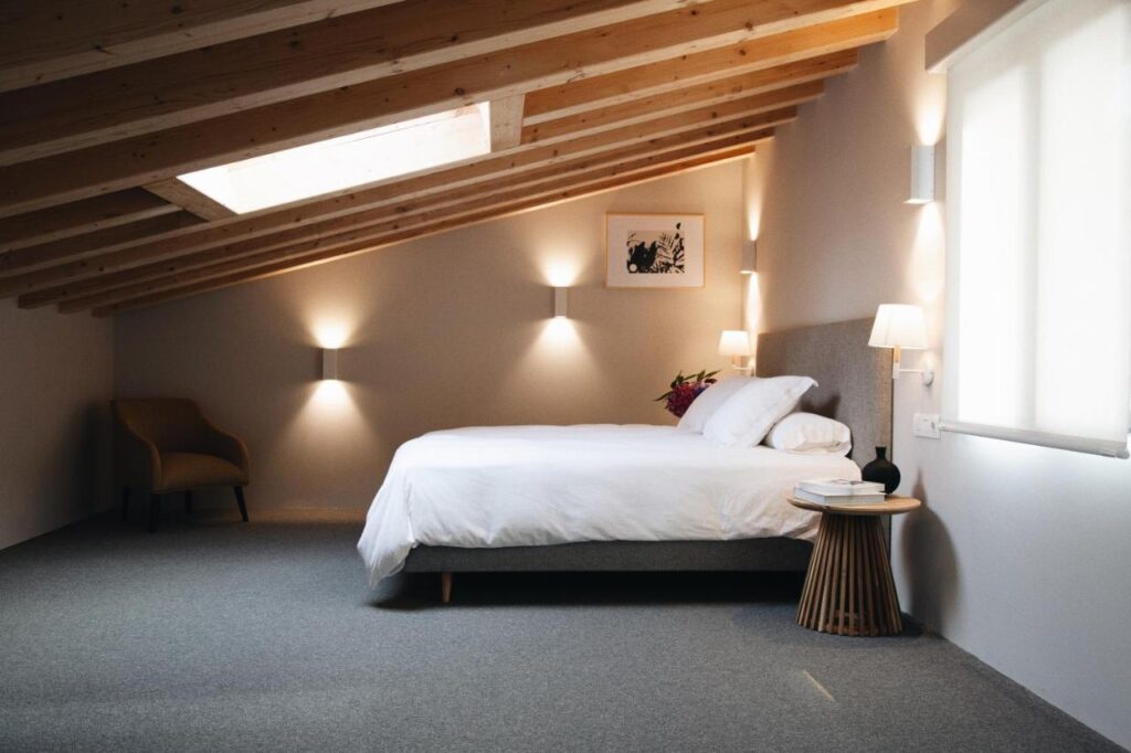 Best Boutique Hotels in San Sebastian 29 A cozy attic bedroom with a sloped wooden ceiling, skylight, and soft lighting—reminiscent of the intimate style found in boutique hotels in San Sebastian—with a neatly made bed, small side table, corner armchair, and minimalist neutral decor.