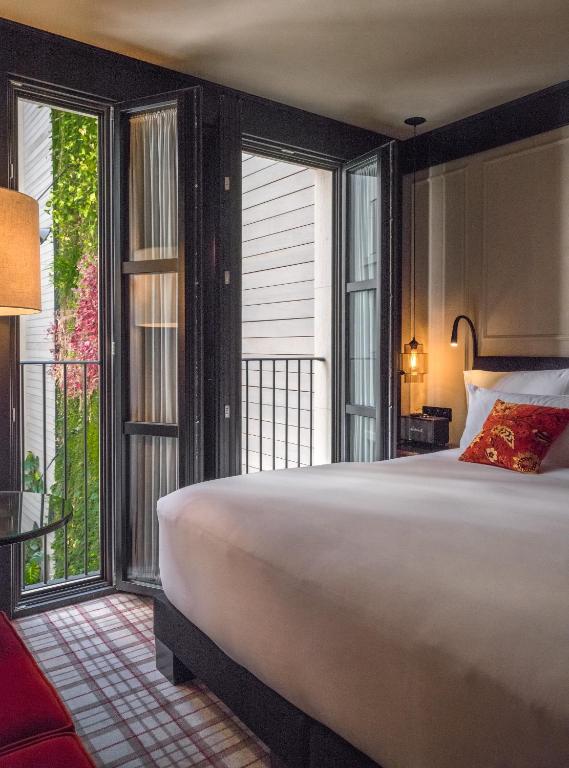 Top Barcelona Hotels with Rooftop Pool 43 A cozy bedroom with a large bed, white linens, red accent pillow, and tall black-framed French windows opening to a green outdoor view—perfect for relaxing after exploring Barcelona hotels with rooftop pool options nearby.