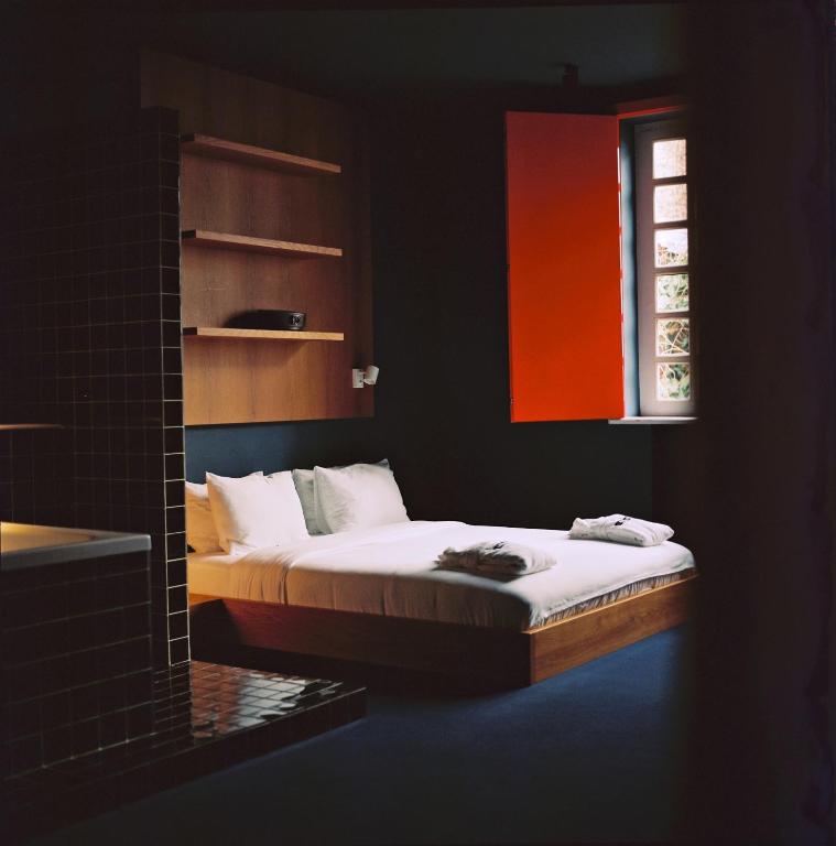 11 Unique Hotels in Georgia You Can't Miss! 10 A minimalist bedroom in one of the unique hotels in Georgia, featuring a low platform bed, white bedding, folded towels, dark walls, open wooden shelves, a tiled partition, and a window with one bright red shutter letting in daylight.