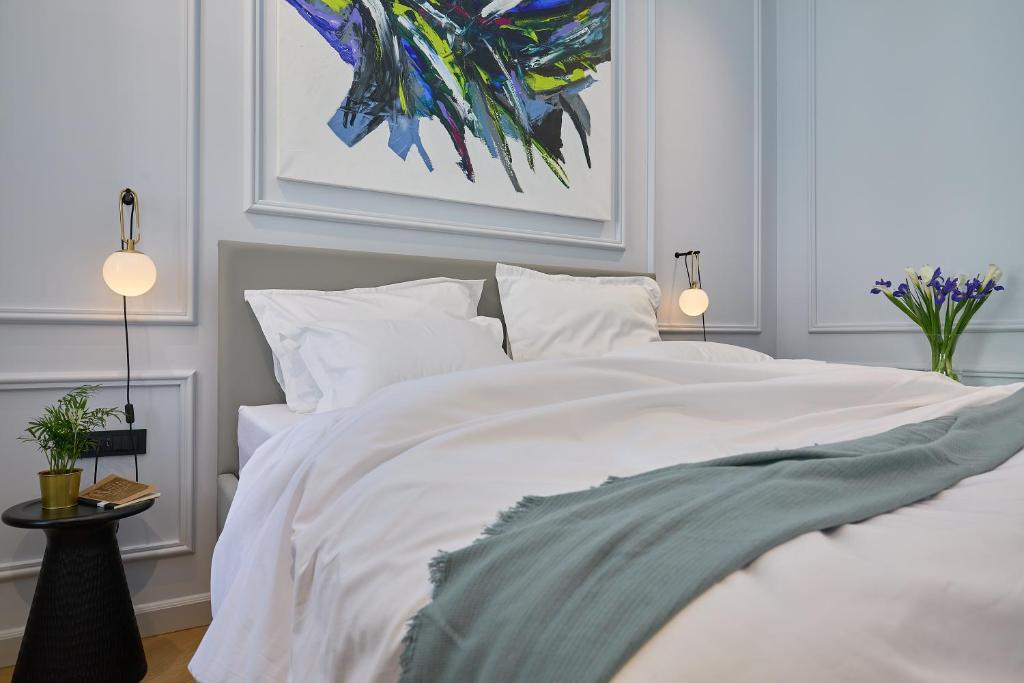 Most Unique Boutique Hotels in Dubrovnik 22 A neatly made bed with white bedding and a light blue throw sits against a light-colored wall, echoing the serene elegance of boutique hotels in Dubrovnik. Abstract artwork hangs above, with wall-mounted lamps and a table topped with a plant and flowers beside the bed.