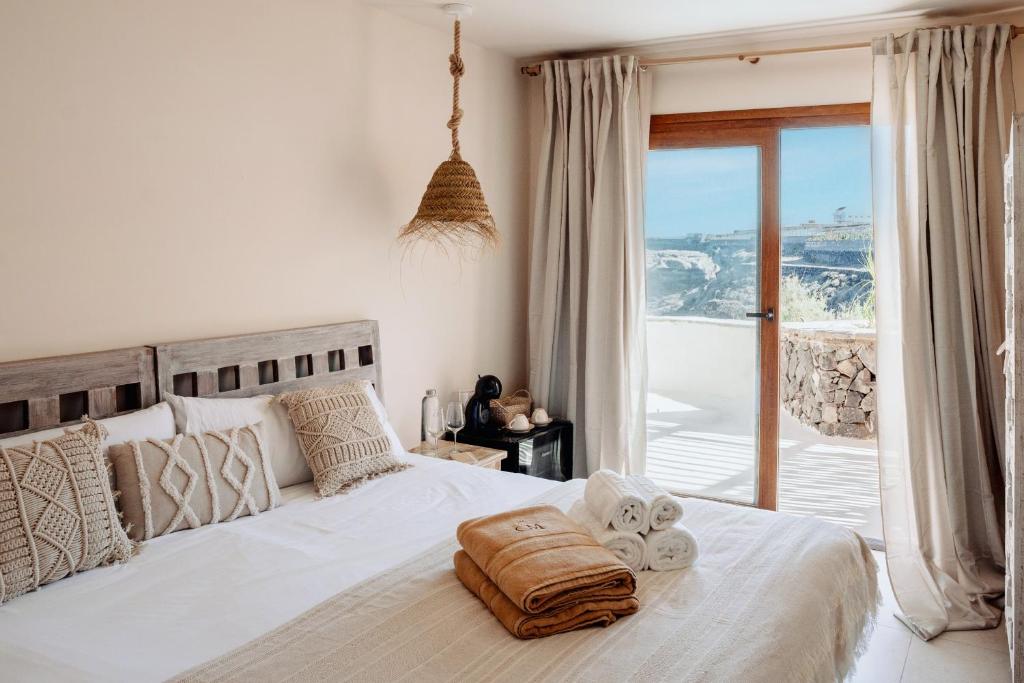 11 UNIQUE Boutique Hotels in Tenerife 22 A cozy, sunlit bedroom in the style of boutique hotels in Tenerife features a double bed with neutral-toned bedding, woven pillows, and a hanging lamp. Towels rest on the bed, while glass doors open to a balcony with a scenic outdoor view.