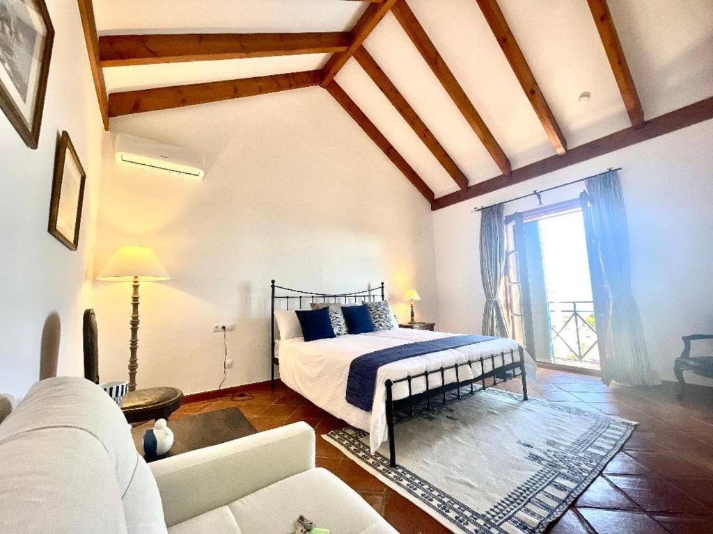 Adults-Only Hotels in Costa del Sol 22 A bright bedroom in one of the top adult only hotels in Costa del Sol, featuring a vaulted wooden beam ceiling, a large bed with white and blue bedding, bedside tables with lamps, a sofa, and a balcony door letting in natural light.