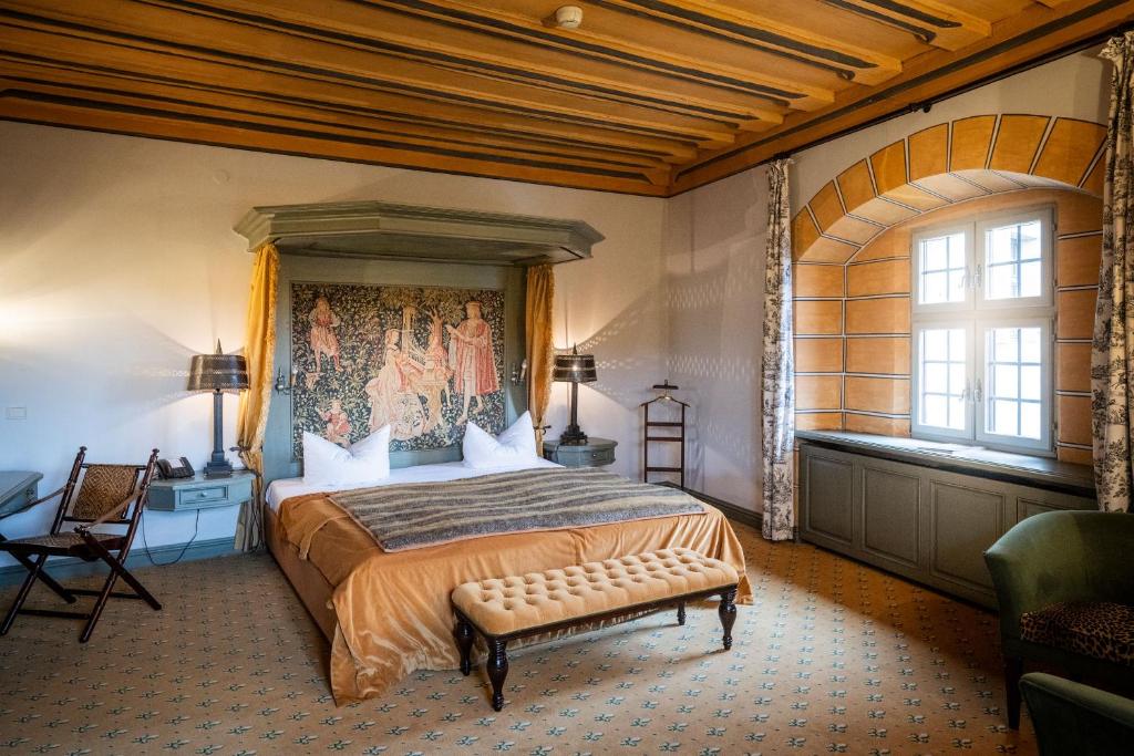 FAIRYTALE Castle Hotels in Bavaria 12 A spacious, elegant bedroom in one of the finest castle hotels in Bavaria, featuring a large bed with a tapestry headboard, golden bedspread, and classic furniture beneath wooden ceiling beams and tall windows.