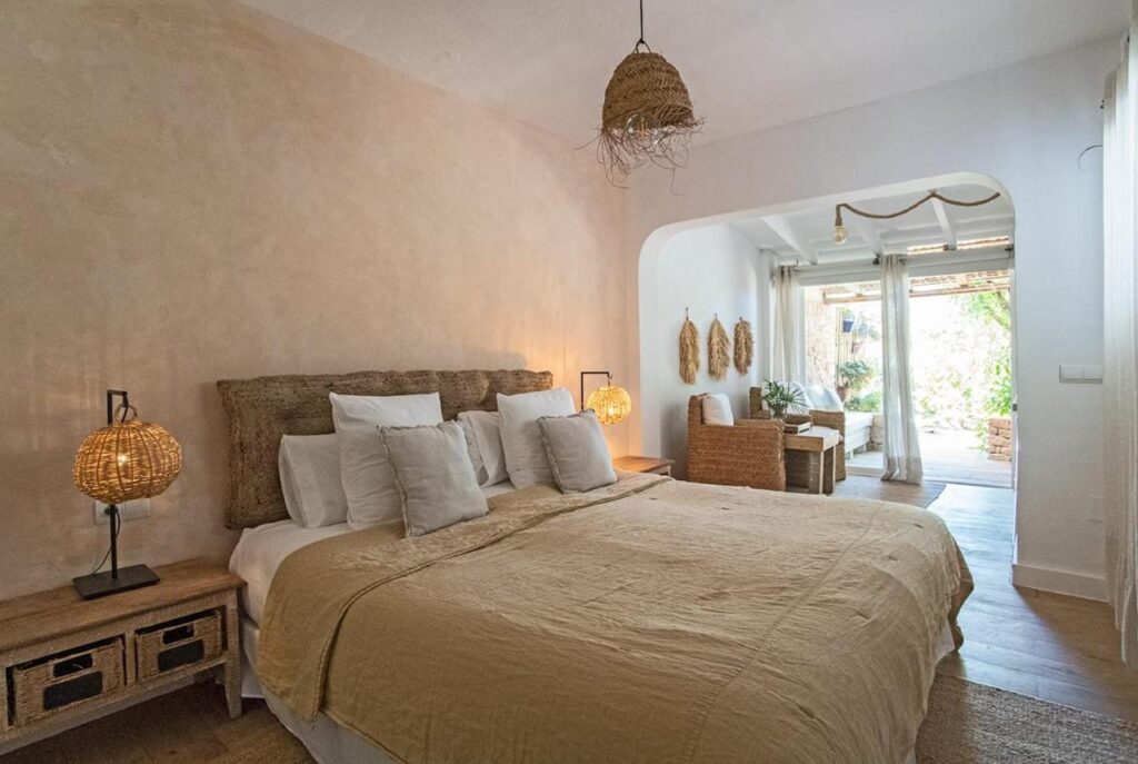A cozy bedroom with a large bed, neutral bedding, wicker lamps, and natural wooden furniture evokes the relaxed style of boutique hotels in Formentera. The room opens to a bright sitting area with rattan chairs and a view of a lush outdoor garden.
